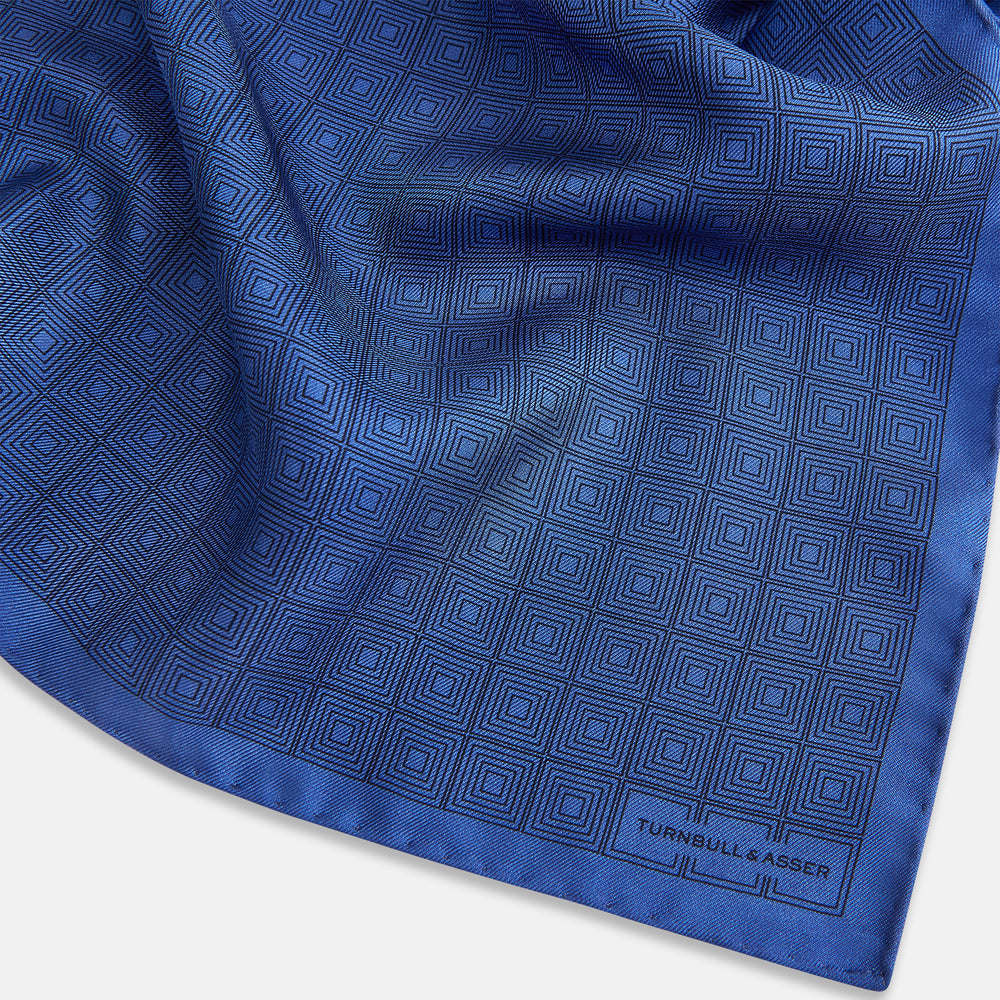 The Geometric Blue Pocket Square in Silk by Turnbull & Asser UK features a vibrant blue design adorned with small, outlined squares arranged in a grid of sharp navy lines. One corner is neatly folded to reveal the reverse side, with "Turnbull & Asser" printed at the bottom right.