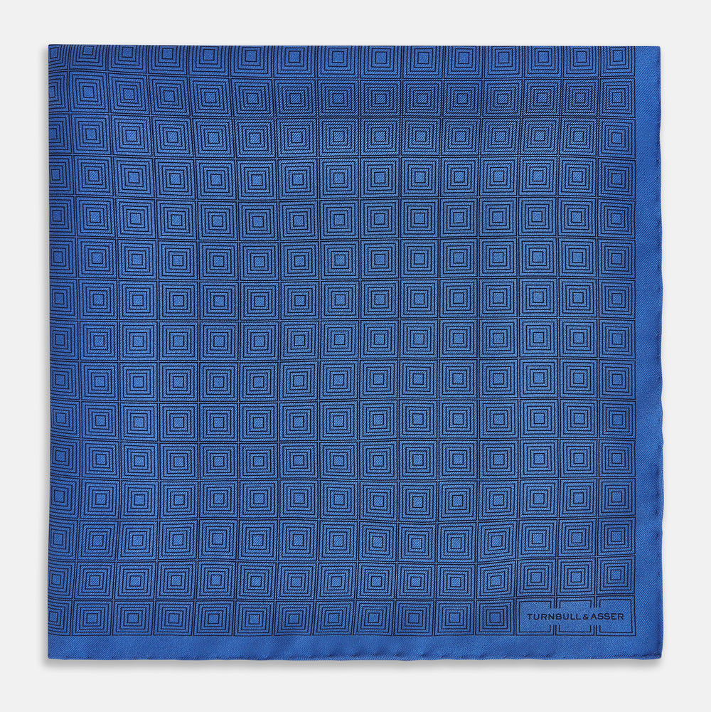 The Geometric Blue Pocket Square in Silk by Turnbull & Asser UK features a vibrant blue design adorned with small, outlined squares arranged in a grid of sharp navy lines. One corner is neatly folded to reveal the reverse side, with "Turnbull & Asser" printed at the bottom right.
