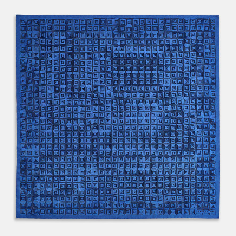 The Geometric Blue Pocket Square in Silk by Turnbull & Asser UK features a vibrant blue design adorned with small, outlined squares arranged in a grid of sharp navy lines. One corner is neatly folded to reveal the reverse side, with "Turnbull & Asser" printed at the bottom right.