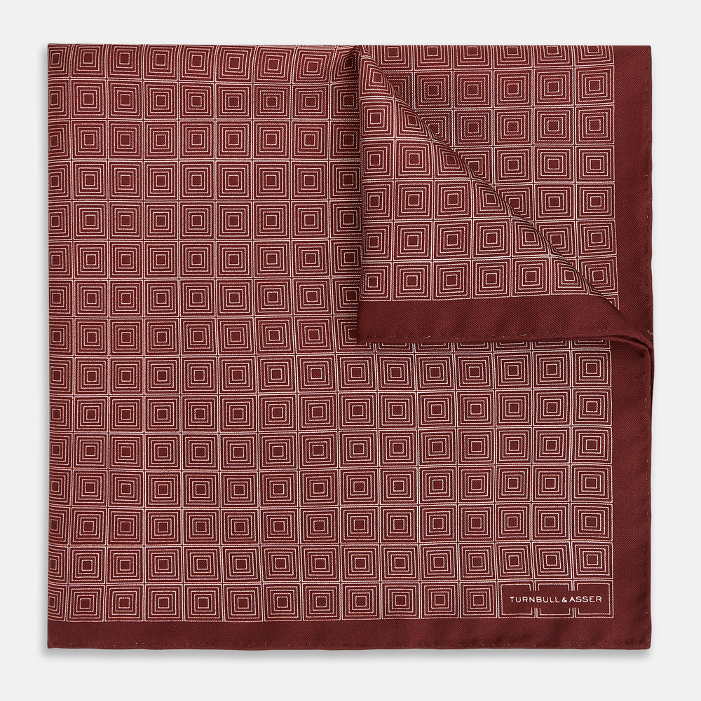 The Geometric Burgundy Pocket Square in Silk by Turnbull & Asser UK features a refined pattern of small, repeating white squares on a rich burgundy background—an ideal accessory for introducing sophistication to any outfit. One corner is artfully folded over to showcase the reverse side of this elegant geometric design, and the "Turnbull & Asser" label appears in the bottom right corner.
