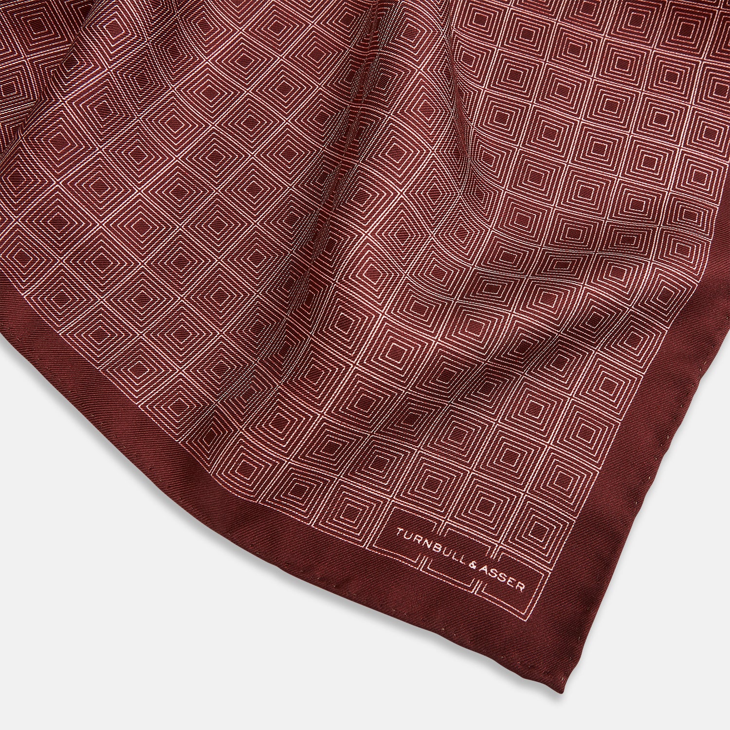 View 2 of product The Geometric Burgundy Pocket Square in Silk by Turnbull & Asser UK features a refined pattern of small, repeating white squares on a rich burgundy background—an ideal accessory for introducing sophistication to any outfit. One corner is artfully folded over to showcase the reverse side of this elegant geometric design, and the "Turnbull & Asser" label appears in the bottom right corner.