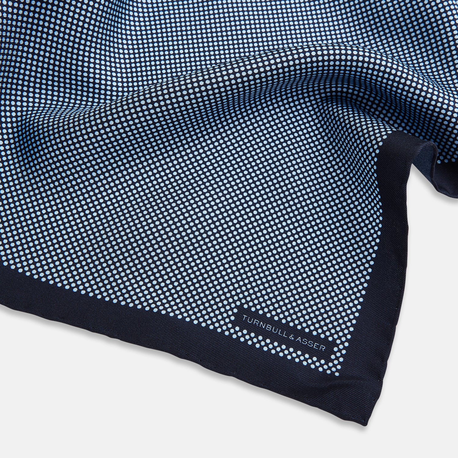 View 2 of product Introducing the Polka Dot Navy Pocket Square in Silk by Turnbull & Asser UK—this elegant accessory features a refined dot motif with small white polka dots and a solid navy border. One corner is artfully folded to reveal the underside, adding visual interest, while the Turnbull & Asser branding is printed in white on one corner, making it an ideal choice for formal tailoring.
