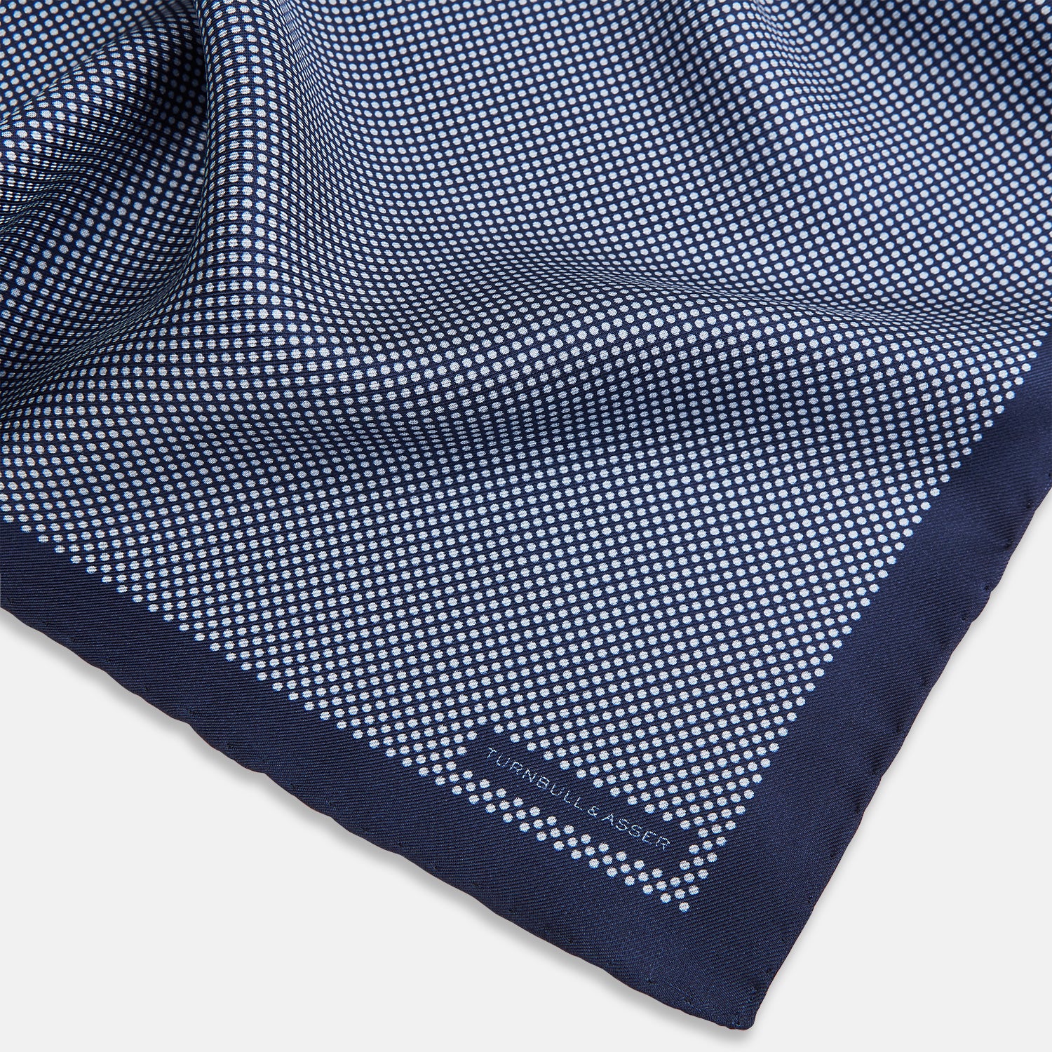 View 2 of product Crafted from silk, the Turnbull & Asser UK Polka dot Denim Pocket Square features a sophisticated navy blue hue adorned with small white polka dots and a solid border. Neatly folded with the brand label visible in one corner, this elegant accessory is ideal for enhancing formal tailoring.
