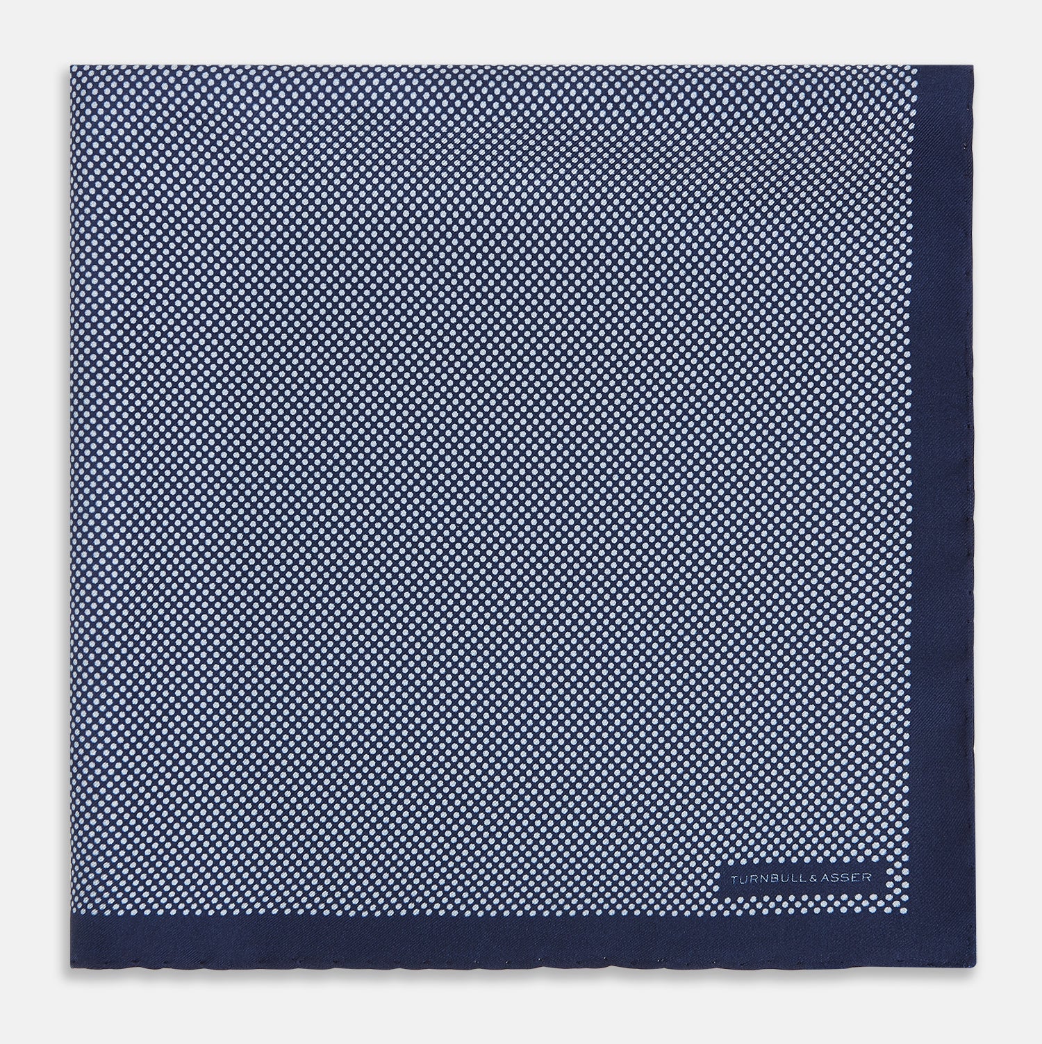 View 3 of product Crafted from silk, the Turnbull & Asser UK Polka dot Denim Pocket Square features a sophisticated navy blue hue adorned with small white polka dots and a solid border. Neatly folded with the brand label visible in one corner, this elegant accessory is ideal for enhancing formal tailoring.