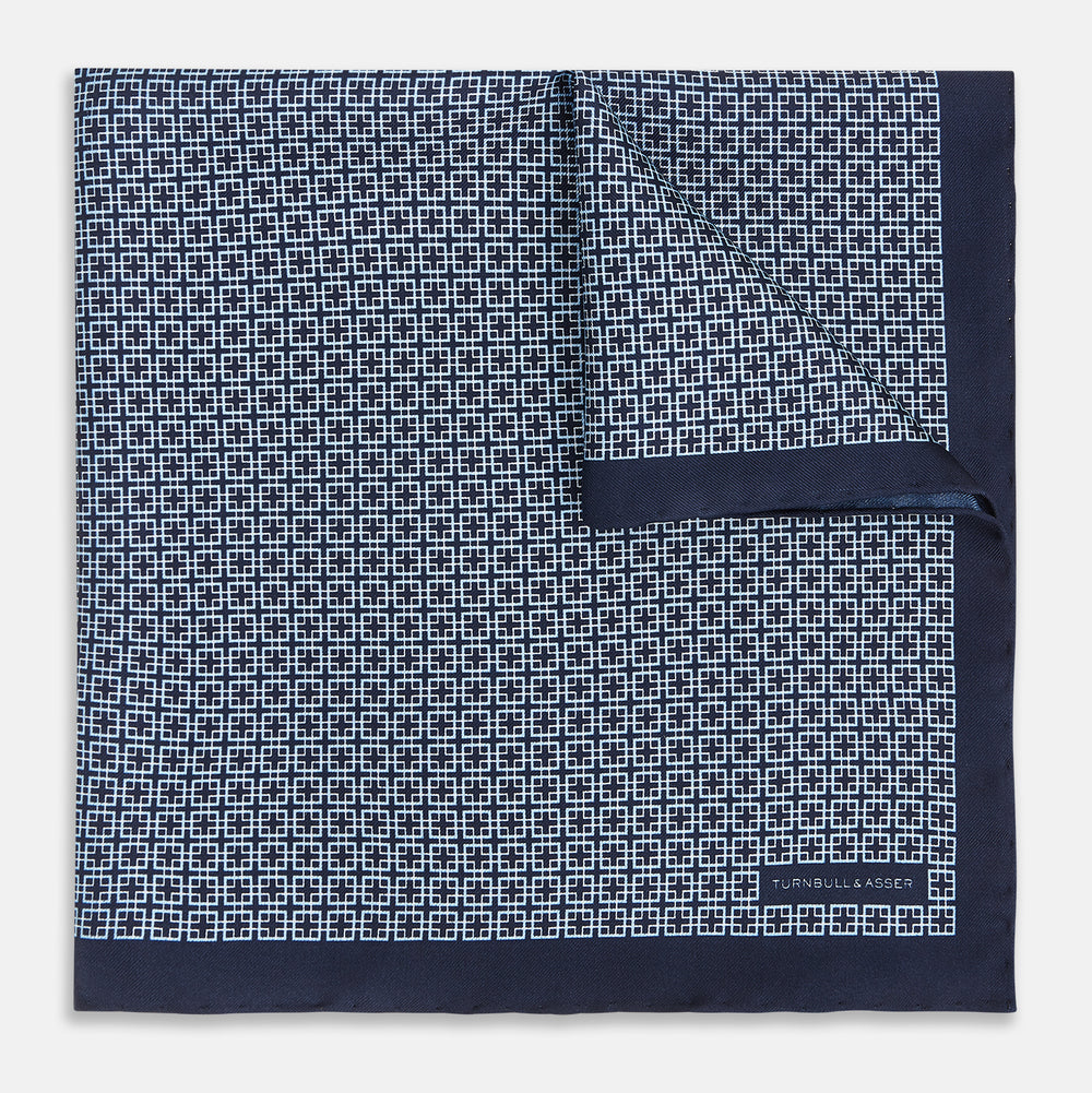 Introducing the Geometric Navy Pocket Square in Silk by Turnbull & Asser UK. This sophisticated accessory showcases a small, repeating geometric pattern in light blue and white on a rich navy background. The pocket square features a solid navy border, while the folded corner reveals the underside adorned with the Turnbull & Asser logo—an elegant finishing touch for any formal ensemble.