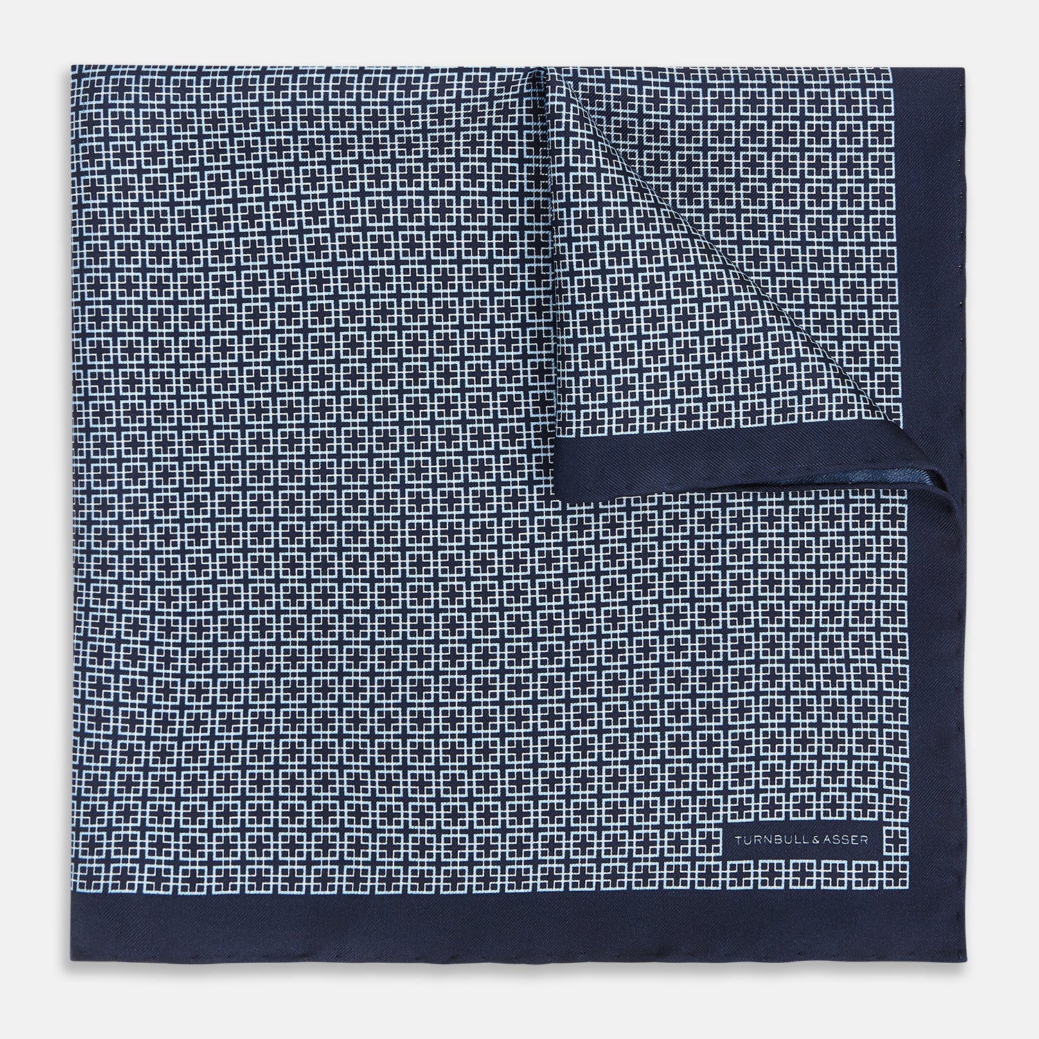 View 1 of product Introducing the Geometric Navy Pocket Square in Silk by Turnbull & Asser UK. This sophisticated accessory showcases a small, repeating geometric pattern in light blue and white on a rich navy background. The pocket square features a solid navy border, while the folded corner reveals the underside adorned with the Turnbull & Asser logo—an elegant finishing touch for any formal ensemble.