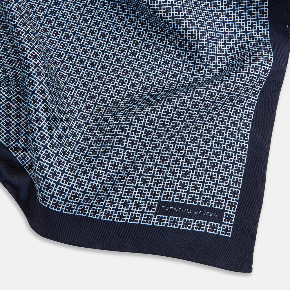 Introducing the Geometric Navy Pocket Square in Silk by Turnbull & Asser UK. This sophisticated accessory showcases a small, repeating geometric pattern in light blue and white on a rich navy background. The pocket square features a solid navy border, while the folded corner reveals the underside adorned with the Turnbull & Asser logo—an elegant finishing touch for any formal ensemble.