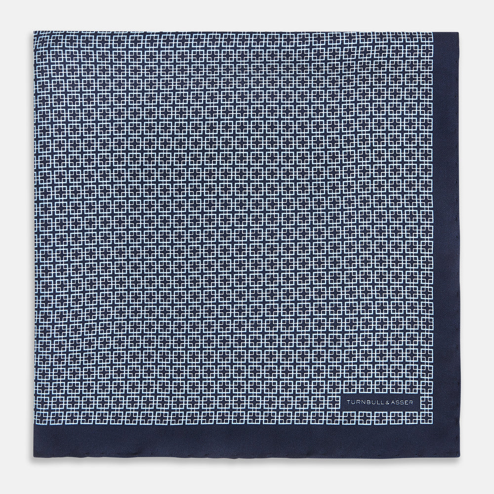 Introducing the Geometric Navy Pocket Square in Silk by Turnbull & Asser UK. This sophisticated accessory showcases a small, repeating geometric pattern in light blue and white on a rich navy background. The pocket square features a solid navy border, while the folded corner reveals the underside adorned with the Turnbull & Asser logo—an elegant finishing touch for any formal ensemble.
