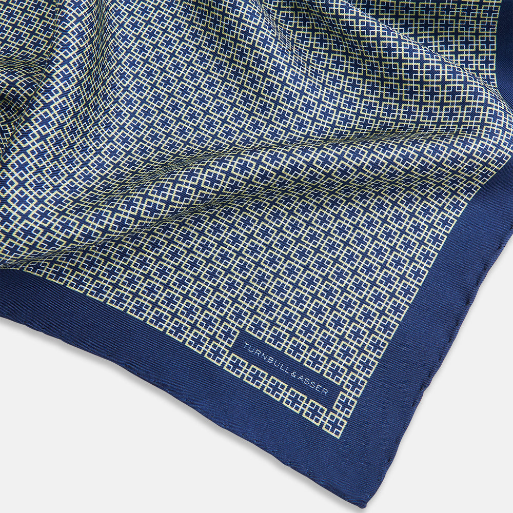 A blue pocket square from Turnbull & Asser UK, featuring a geometric white square pattern and a solid blue border. One corner is folded to highlight the striking design on both sides, with the brand name displayed on the bottom right. Product name: POCKET SQUARE SQUARE PATTERNED BANANA.