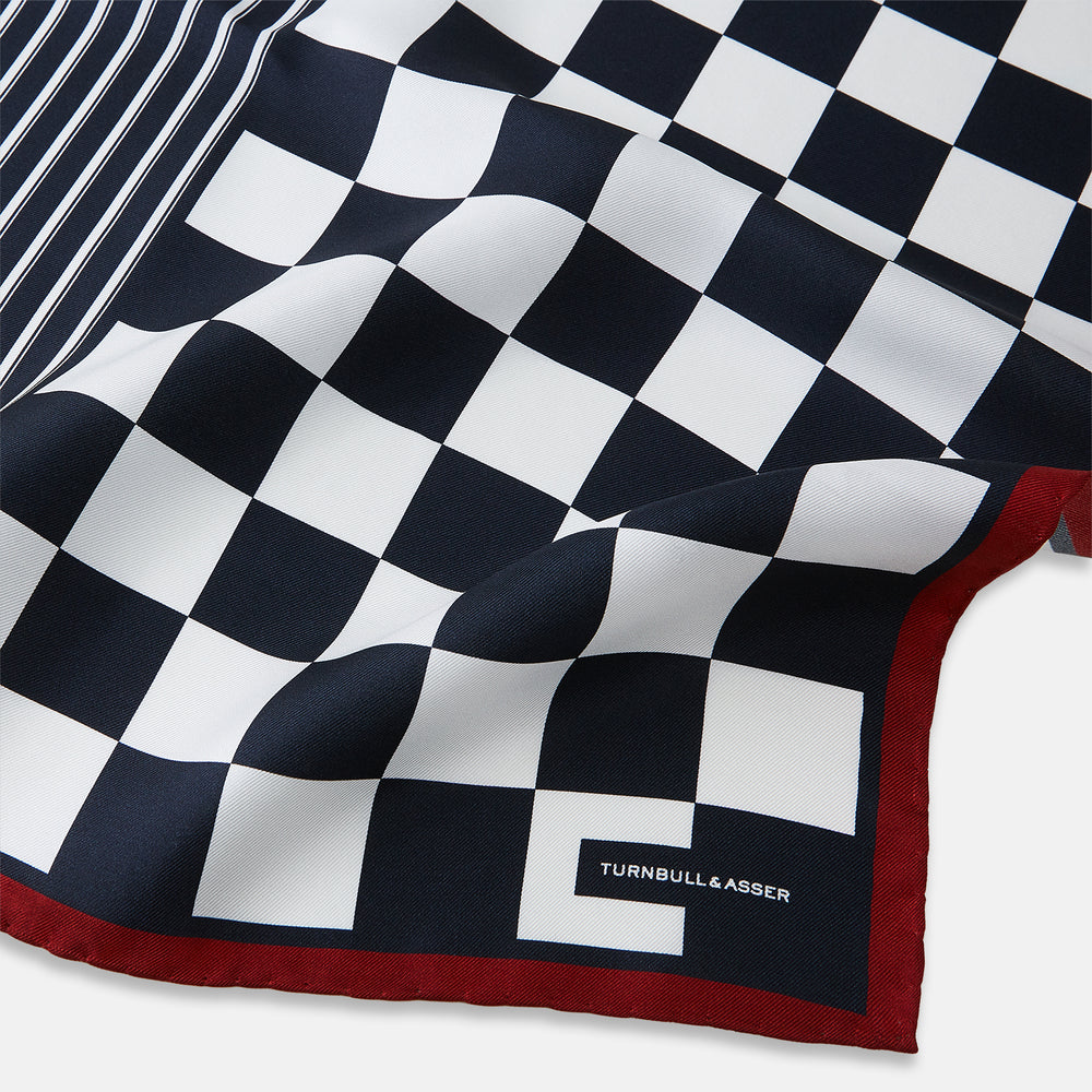 Introducing the Chequered Navy on Red Frame Pocket Square in Silk from Turnbull & Asser UK. This limited edition accessory, part of the Driver’s Capsule Collection, features a bold navy and white checkerboard pattern bordered by deep red silk. The bottom right corner is subtly detailed with white text reading “Turnbull & Asser” and “2 of 20.”.