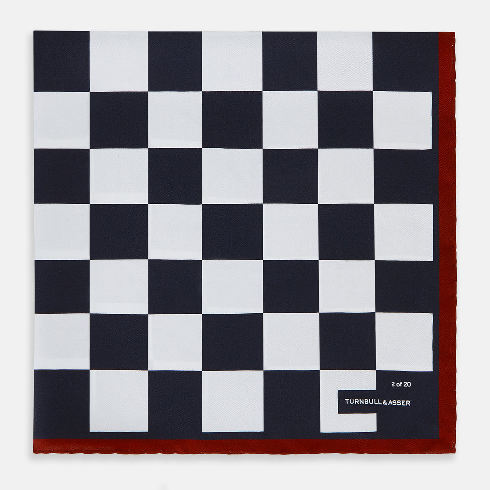Introducing the Chequered Navy on Red Frame Pocket Square in Silk from Turnbull & Asser UK. This limited edition accessory, part of the Driver’s Capsule Collection, features a bold navy and white checkerboard pattern bordered by deep red silk. The bottom right corner is subtly detailed with white text reading “Turnbull & Asser” and “2 of 20.”.