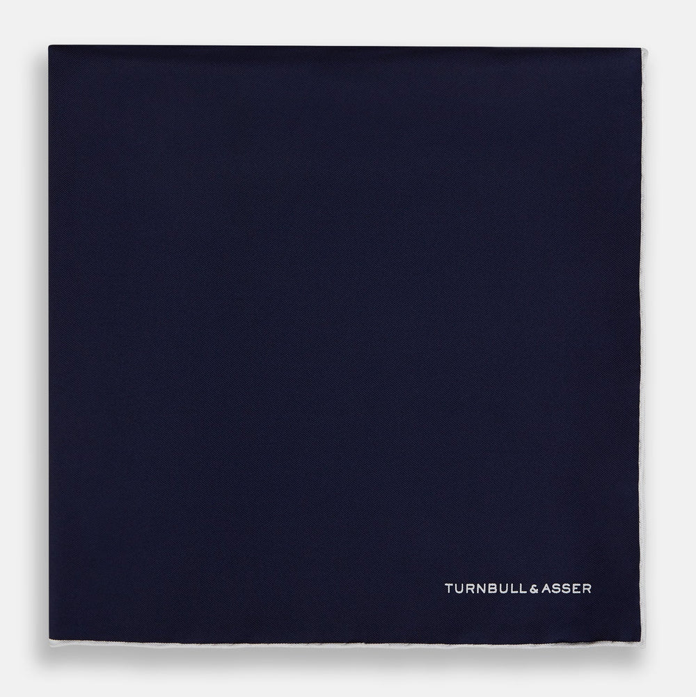The Navy Piped Silk Pocket Square by Turnbull & Asser features white-stitched edges and subtle brand embroidery, ideal for enhancing formal looks. Shown neatly folded on a plain white background.