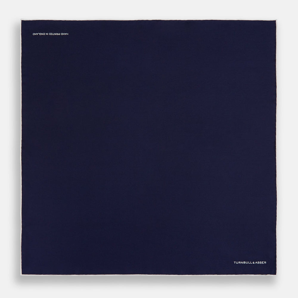 The Navy Piped Silk Pocket Square by Turnbull & Asser features white-stitched edges and subtle brand embroidery, ideal for enhancing formal looks. Shown neatly folded on a plain white background.