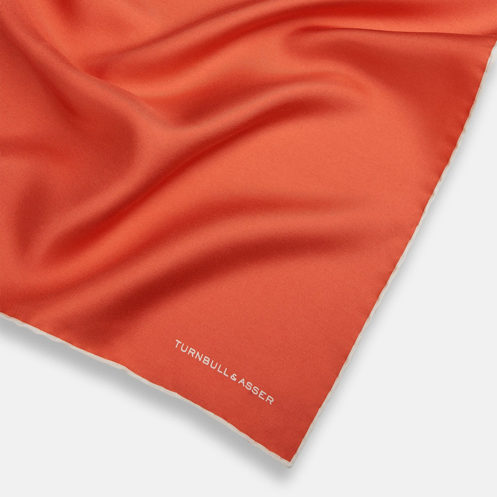 The Orange and White Piped Silk Pocket Square by Turnbull & Asser features a solid orange design with white piping and the brand’s logo in the bottom right corner, making it an elegant addition to any formal outfit.