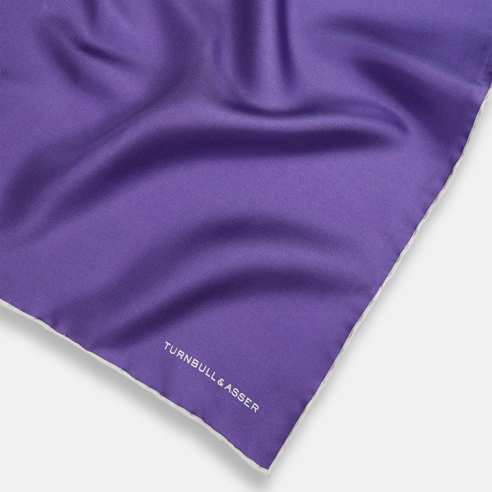 The Turnbull & Asser Violet and Ecru Piped Silk Pocket Square features a stitched edge and white brand logo, making it an elegant addition to formalwear.