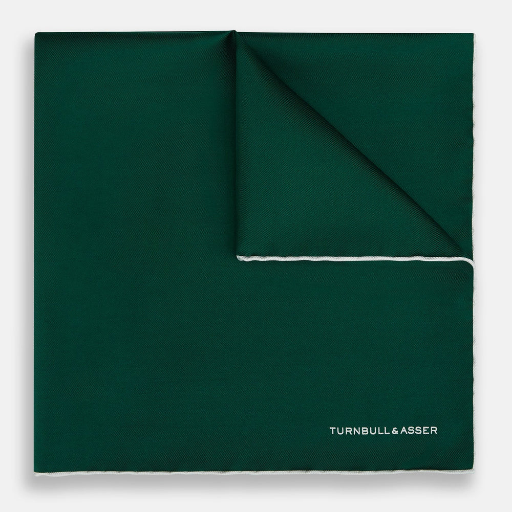 The FOREST GREEN AND ECRU PIPED SILK POCKET SQUARE by Turnbull & Asser features forest green silk with ecru piping, neatly folded to display both sides, and the brand name printed in white in the lower right corner—ideal for formal attire.