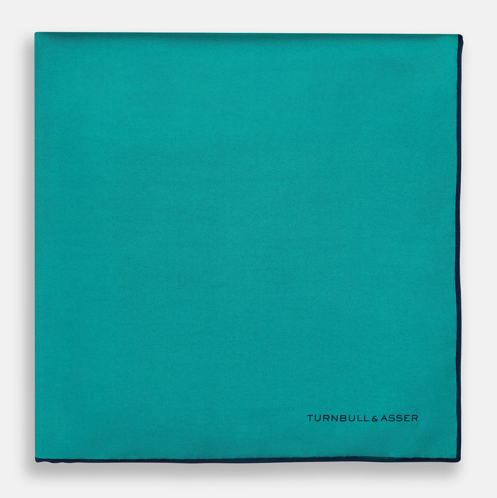 The Turquoise Piped Silk Pocket Square by Turnbull & Asser features dark blue edging and the brand’s logo, offering a sophisticated accent to formal outfits.