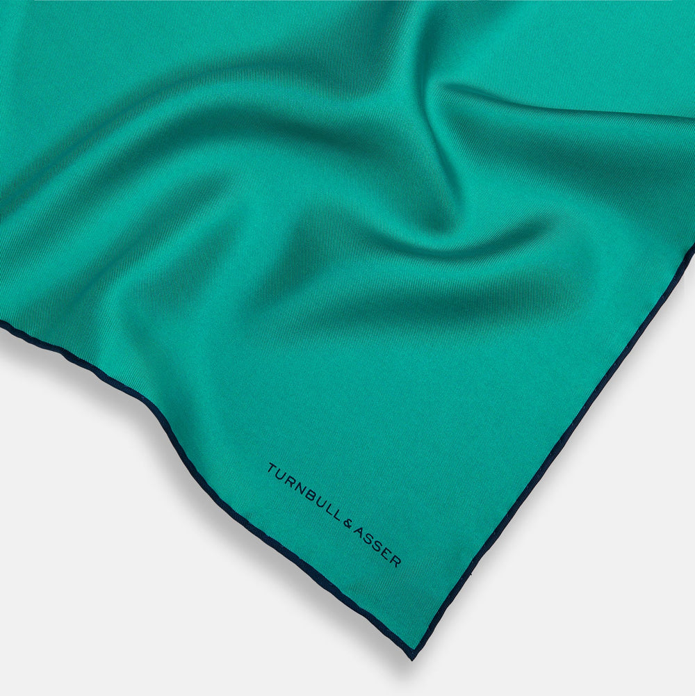 The Turquoise Piped Silk Pocket Square by Turnbull & Asser features dark blue edging and the brand’s logo, offering a sophisticated accent to formal outfits.