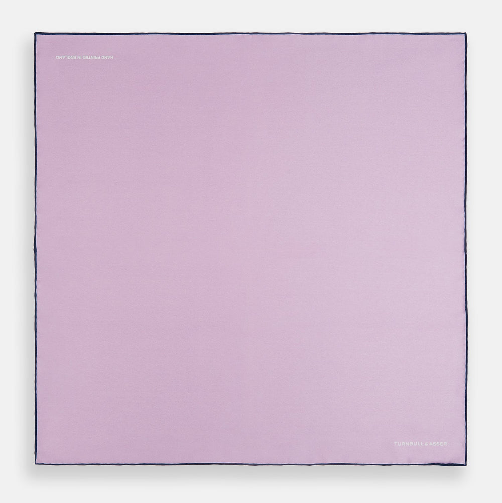 The Turnbull & Asser Lilac Piped Silk Pocket Square features dark edging and a white brand logo in one corner, making it a stylish choice for formal attire. Displayed against a white background.