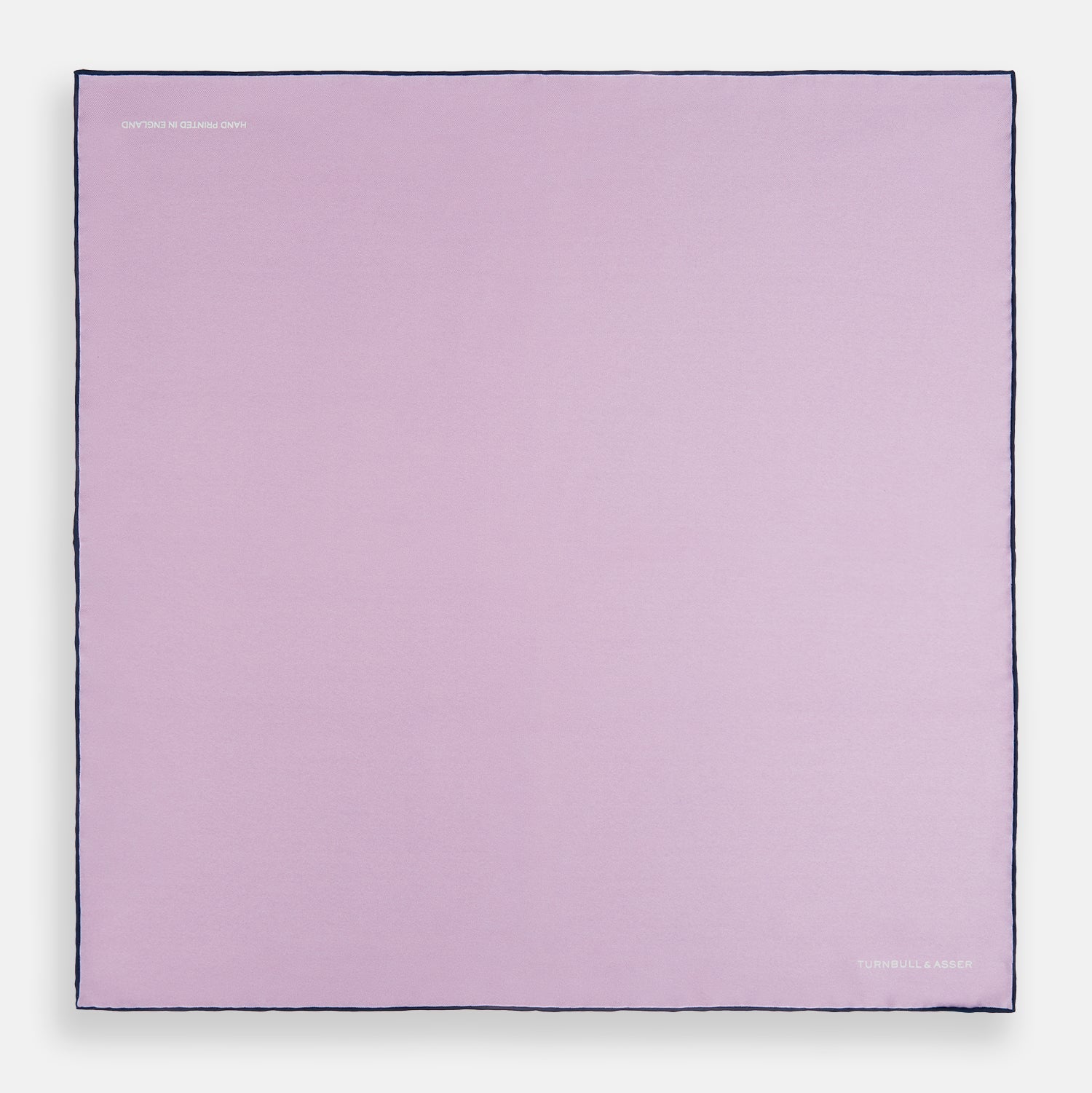 View 4 of product The Turnbull & Asser Lilac Piped Silk Pocket Square features dark edging and a white brand logo in one corner, making it a stylish choice for formal attire. Displayed against a white background.