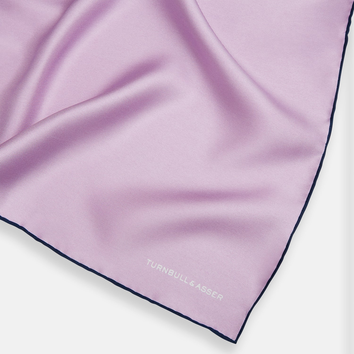 View 2 of product The Turnbull & Asser Lilac Piped Silk Pocket Square features dark edging and a white brand logo in one corner, making it a stylish choice for formal attire. Displayed against a white background.