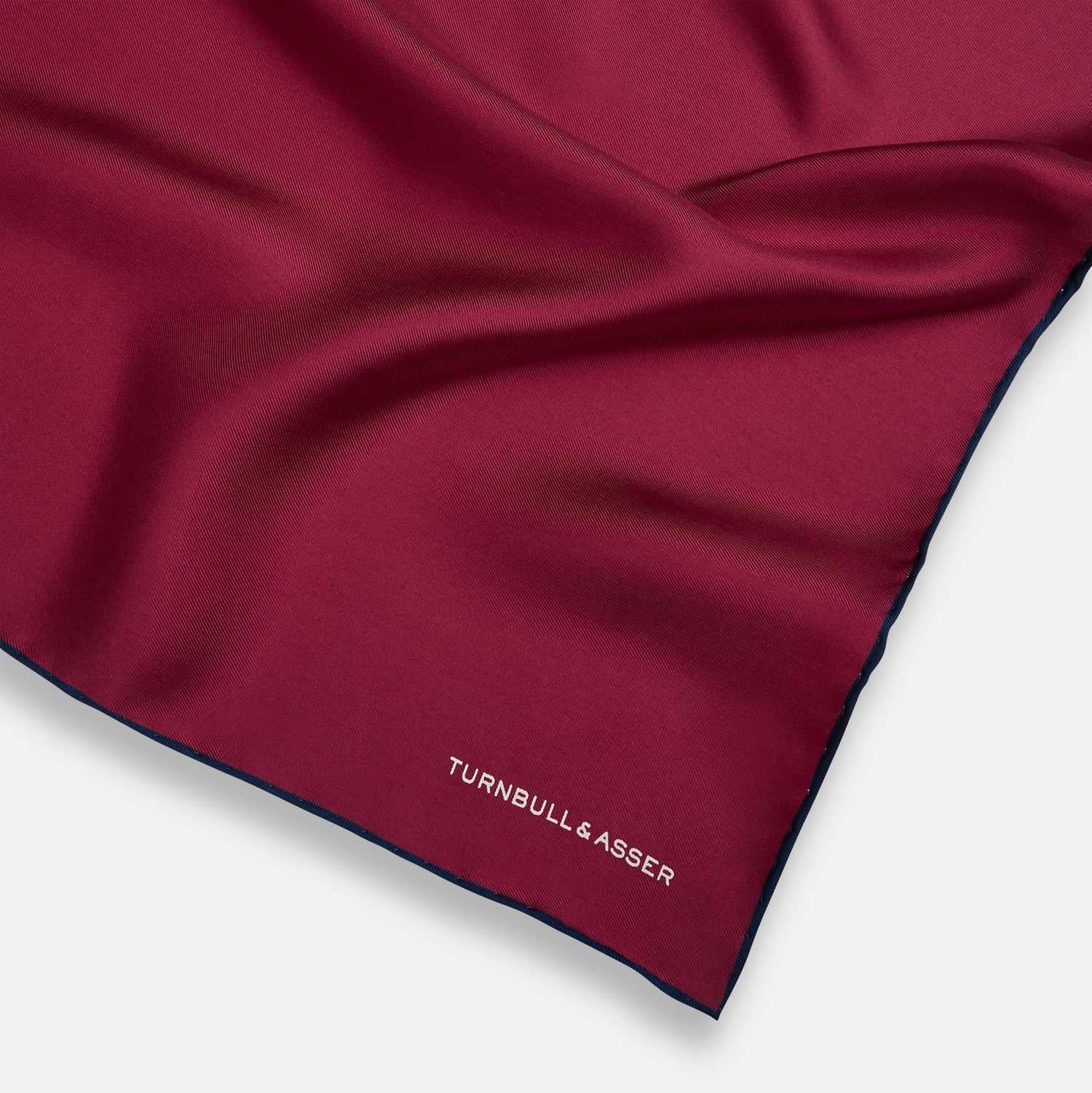 View 2 of product The Mauve Piped Silk Pocket Square by Turnbull & Asser features a dark blue edge and white embroidered logo in the lower right corner, making it ideal for formal occasions. Displayed on a plain white background.