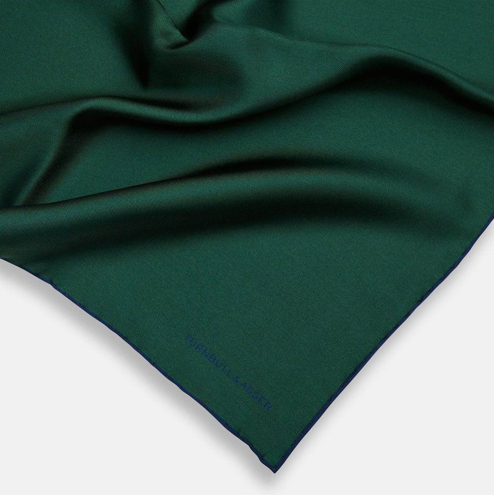 The Turnbull & Asser Petrol Green Piped Silk Pocket Square features a visible stitched edge and a discreet blue logo on the turned-down corner, adding an elegant accent to formal attire.