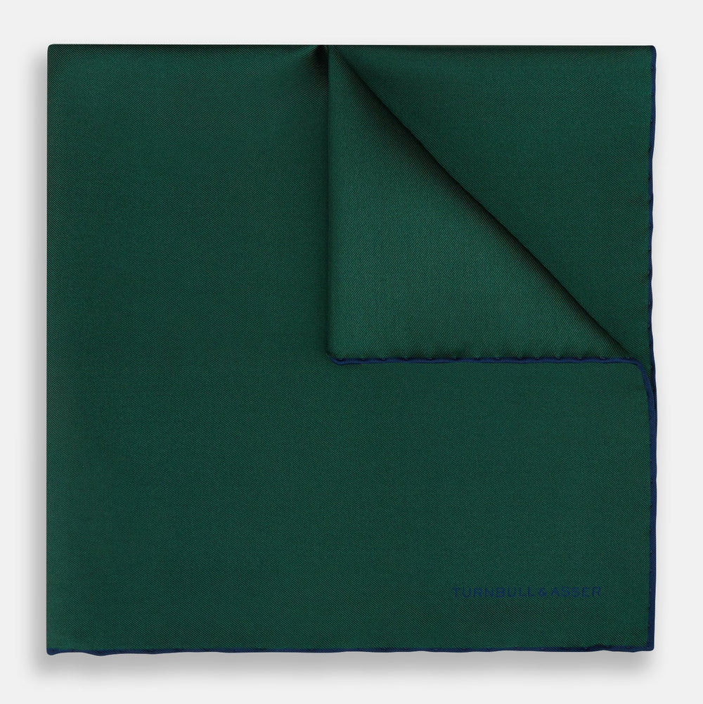 The Turnbull & Asser Petrol Green Piped Silk Pocket Square features a visible stitched edge and a discreet blue logo on the turned-down corner, adding an elegant accent to formal attire.