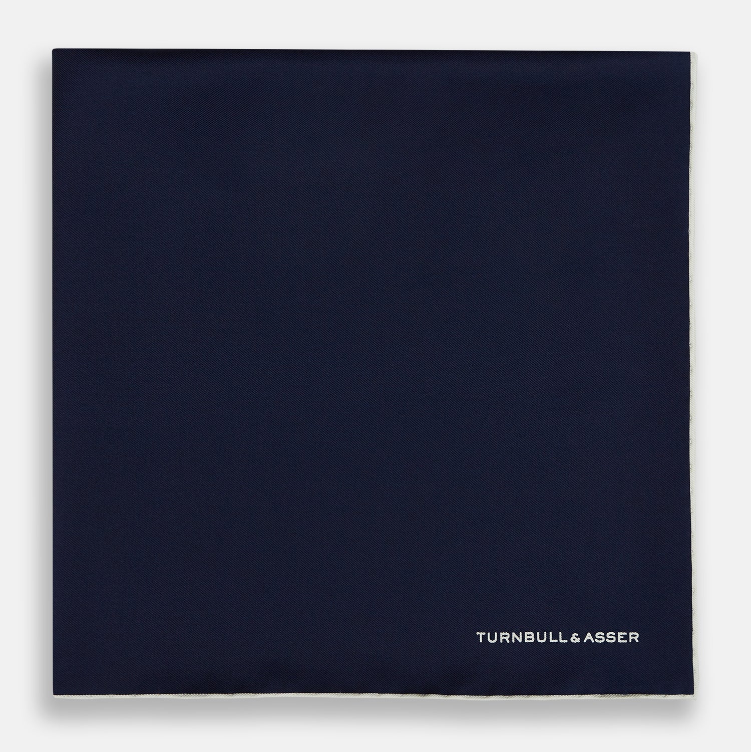 View 3 of product The Dark Blue Piped Silk Pocket Square by Turnbull & Asser features white piping, a one-corner fold revealing the underside, and the brand's name embroidered in white—perfect for formal occasions.