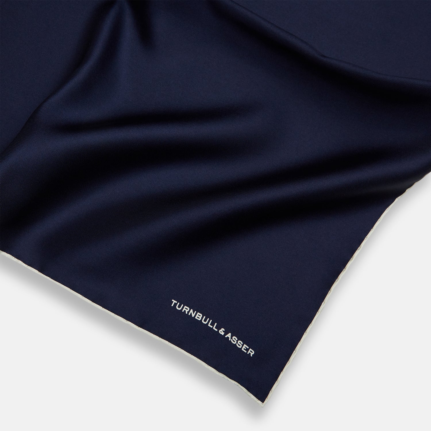 View 2 of product The Dark Blue Piped Silk Pocket Square by Turnbull & Asser features white piping, a one-corner fold revealing the underside, and the brand's name embroidered in white—perfect for formal occasions.