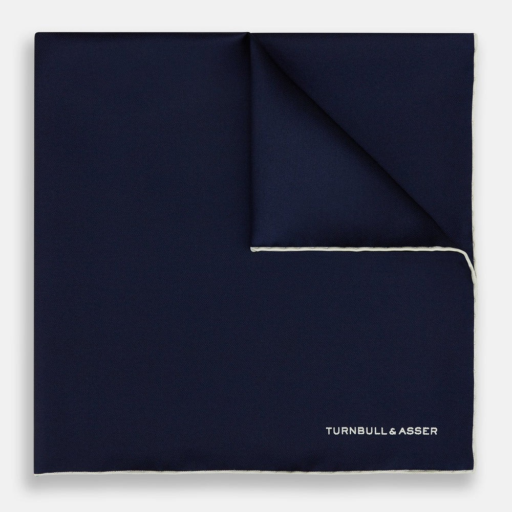The Dark Blue Piped Silk Pocket Square by Turnbull & Asser features white piping, a one-corner fold revealing the underside, and the brand's name embroidered in white—perfect for formal occasions.