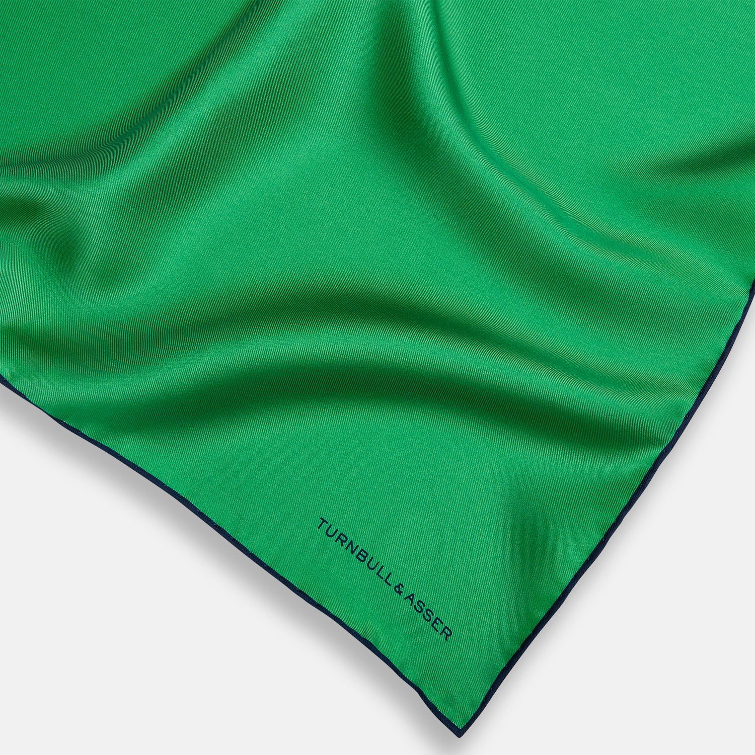 View 2 of product The Green Piped Silk Pocket Square by Turnbull & Asser features dark blue stitched edges and a neat fold—ideal for formal occasions. The Turnbull & Asser name is printed in black on the lower right corner.