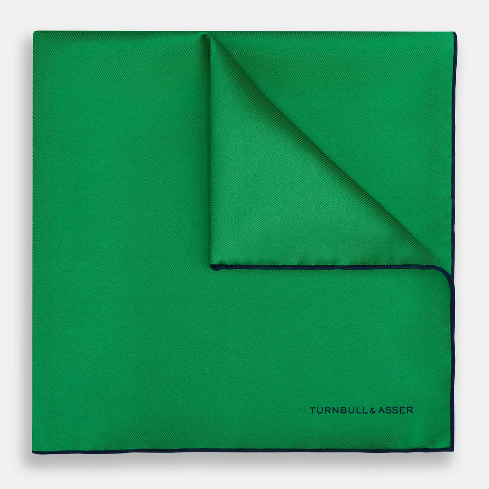The Green Piped Silk Pocket Square by Turnbull & Asser features dark blue stitched edges and a neat fold—ideal for formal occasions. The Turnbull & Asser name is printed in black on the lower right corner.