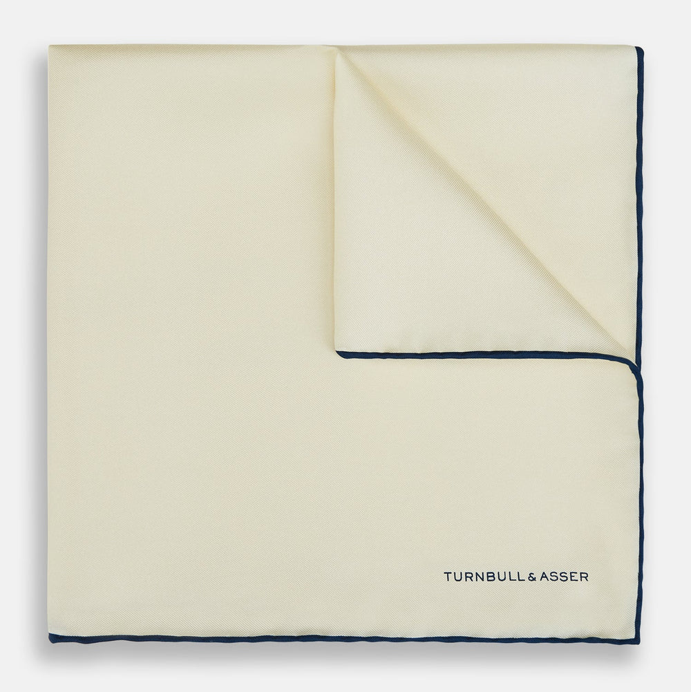 The Ecru and Navy Blue Piped Silk Pocket Square by Turnbull & Asser features contrasting navy piping and finely embroidered branding—an elegant finishing touch to any formal look.