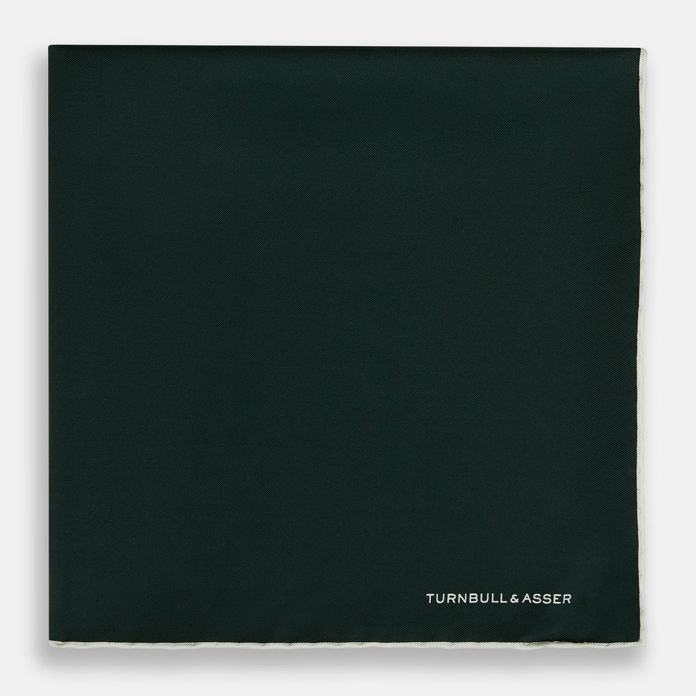 The DARK GREEN AND ECRU PIPED SILK POCKET SQUARE by Turnbull & Asser features ecru edging and the brand's logo in one corner, making it a refined choice for formal attire. One corner is folded to show the reverse side.