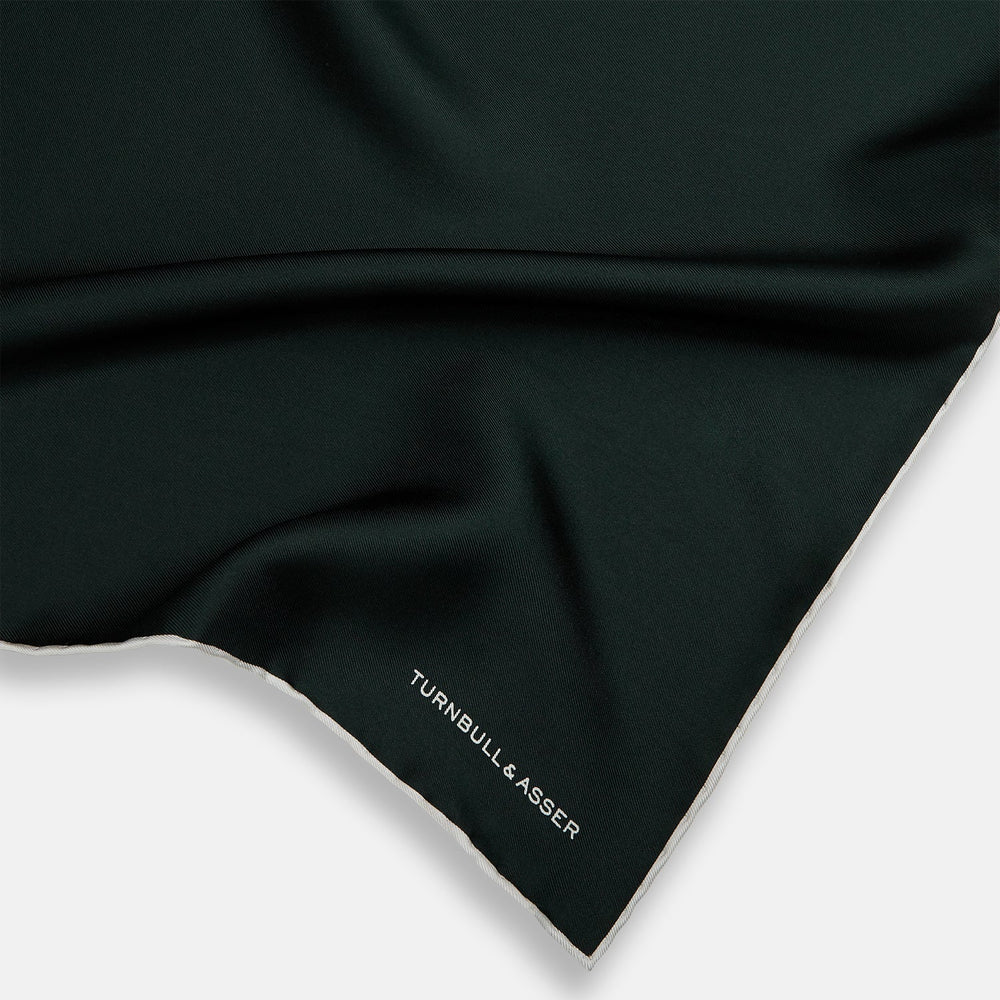 The DARK GREEN AND ECRU PIPED SILK POCKET SQUARE by Turnbull & Asser features ecru edging and the brand's logo in one corner, making it a refined choice for formal attire. One corner is folded to show the reverse side.