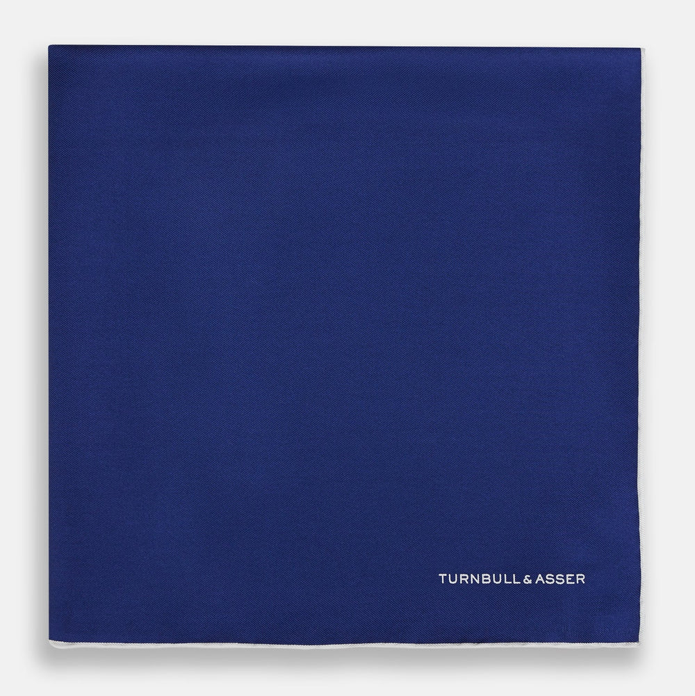 The Turnbull & Asser Royal Blue Piped Silk Pocket Square features white piping and the brand logo embroidered in white at the bottom right, making it perfect for formalwear.