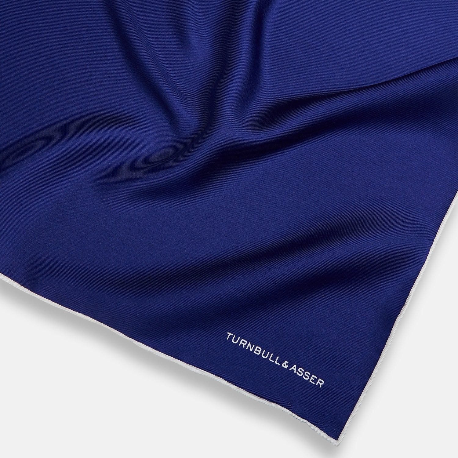 View 2 of product The Turnbull & Asser Royal Blue Piped Silk Pocket Square features white piping and the brand logo embroidered in white at the bottom right, making it perfect for formalwear.