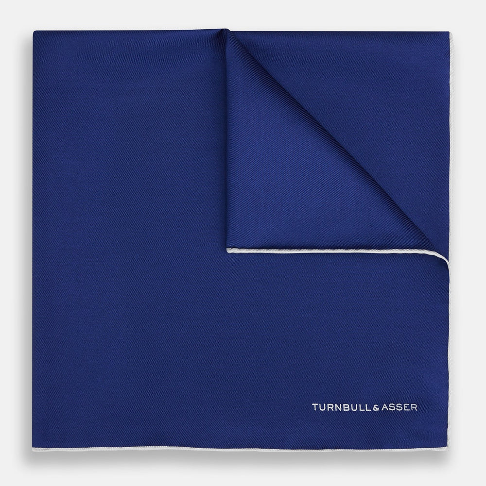 The Turnbull & Asser Royal Blue Piped Silk Pocket Square features white piping and the brand logo embroidered in white at the bottom right, making it perfect for formalwear.