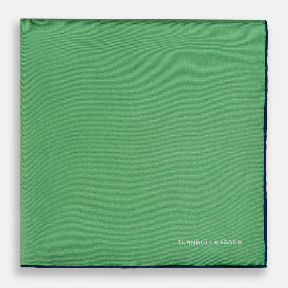 The Fern Green Piped Silk Pocket Square by Turnbull & Asser features a dark blue edge and white embroidered branding at the corner—an elegant accessory for formal wear.