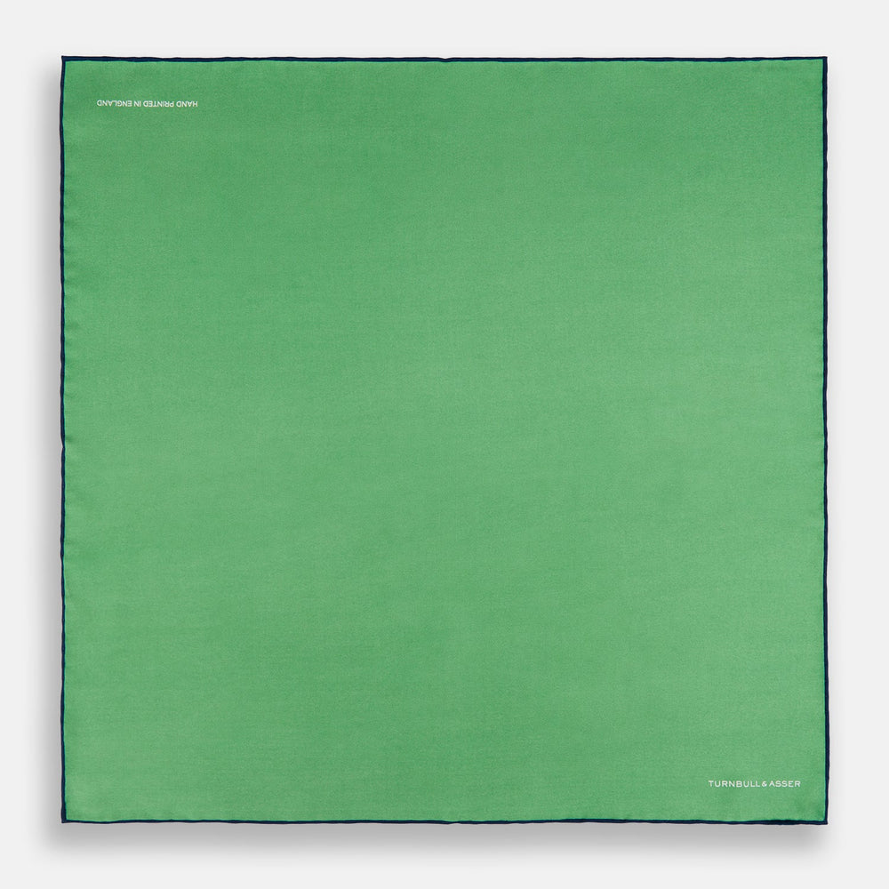 The Fern Green Piped Silk Pocket Square by Turnbull & Asser features a dark blue edge and white embroidered branding at the corner—an elegant accessory for formal wear.