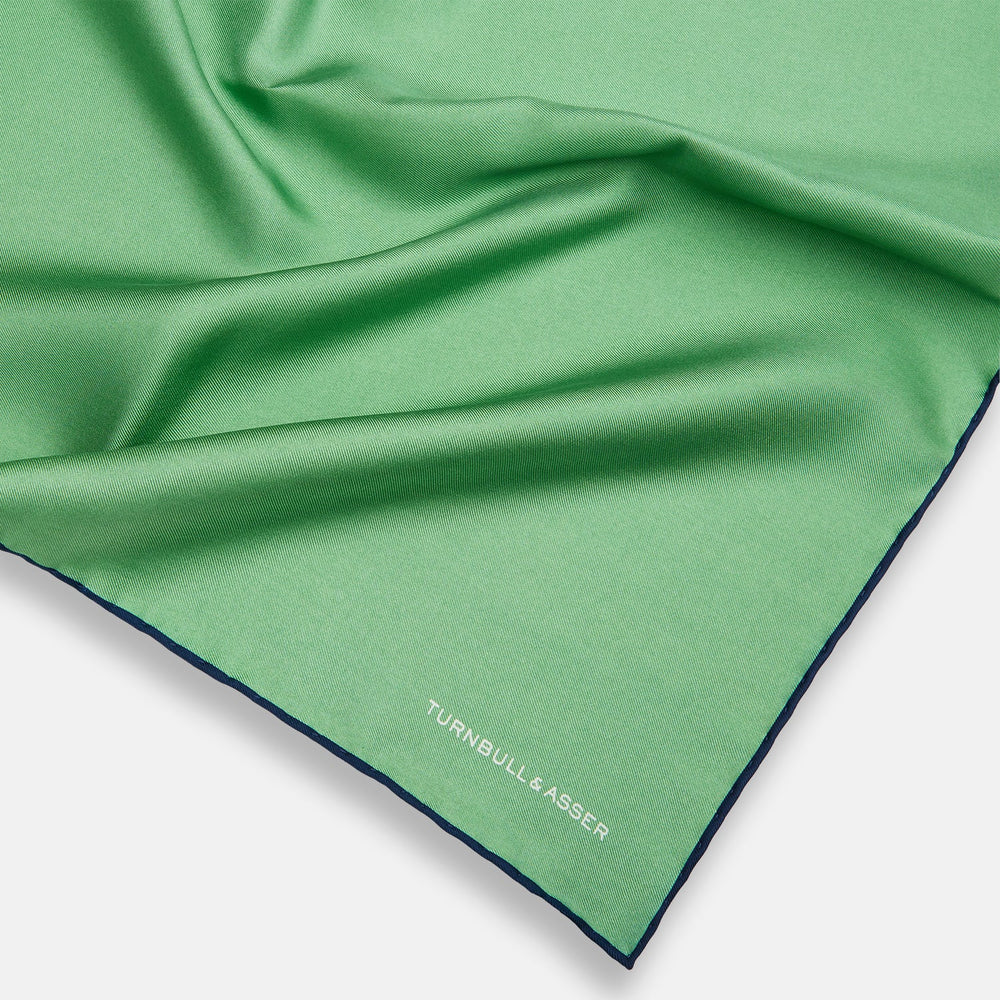 The Fern Green Piped Silk Pocket Square by Turnbull & Asser features a dark blue edge and white embroidered branding at the corner—an elegant accessory for formal wear.