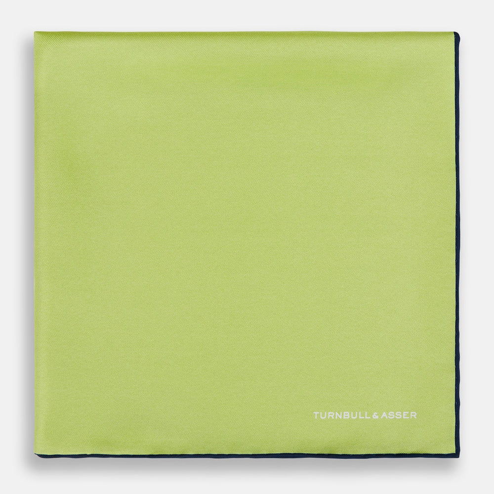 The Pistachio Green and Navy Piped Silk Pocket Square by Turnbull & Asser features crisp navy edging and an elegant brand logo, making it a refined addition to any formal outfit.