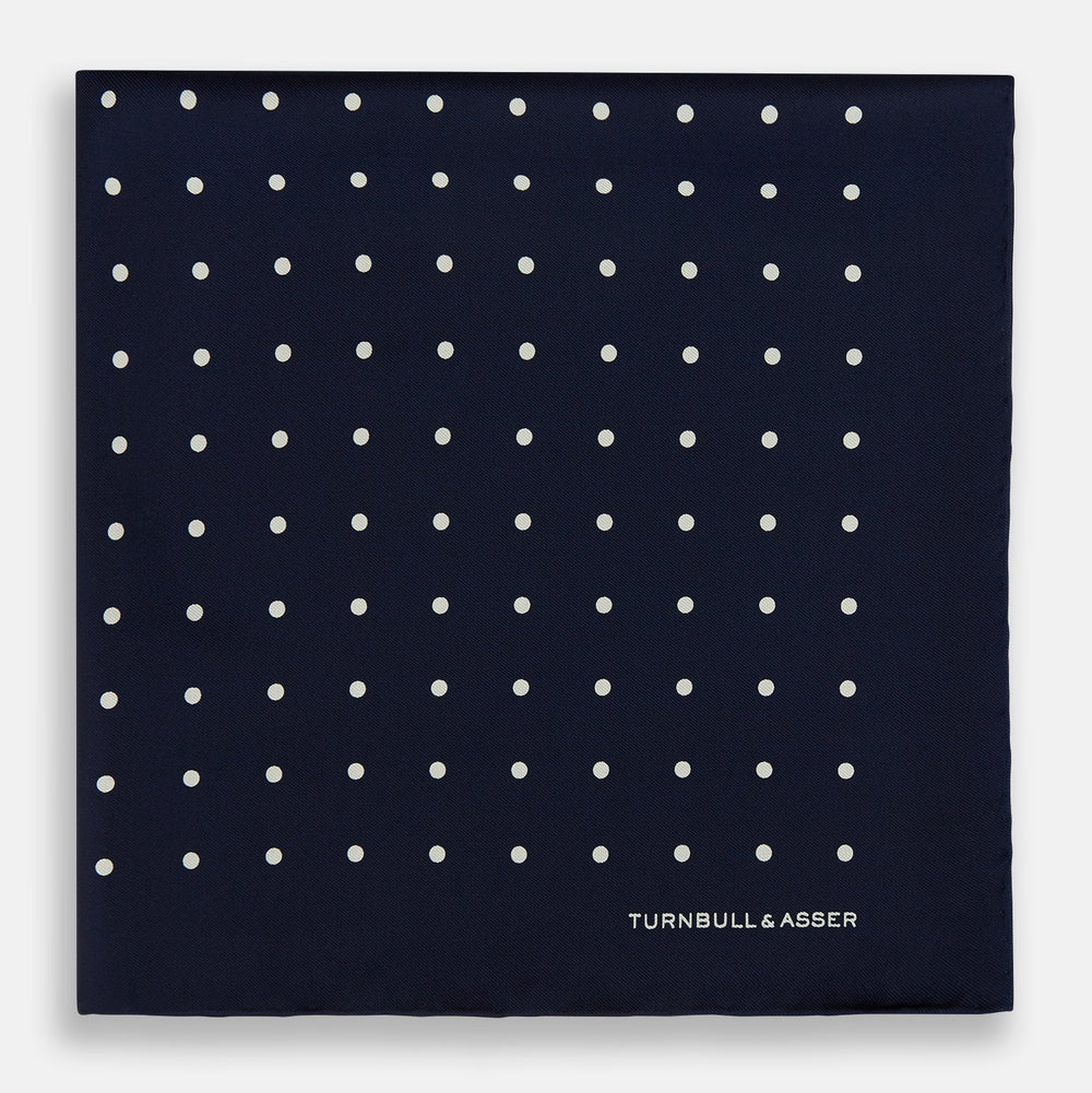 The Turnbull & Asser Navy and White Spot Silk Pocket Square features evenly spaced white polka dots and the brand logo in one corner—a stylish finishing touch for formal looks. One corner is neatly folded over.