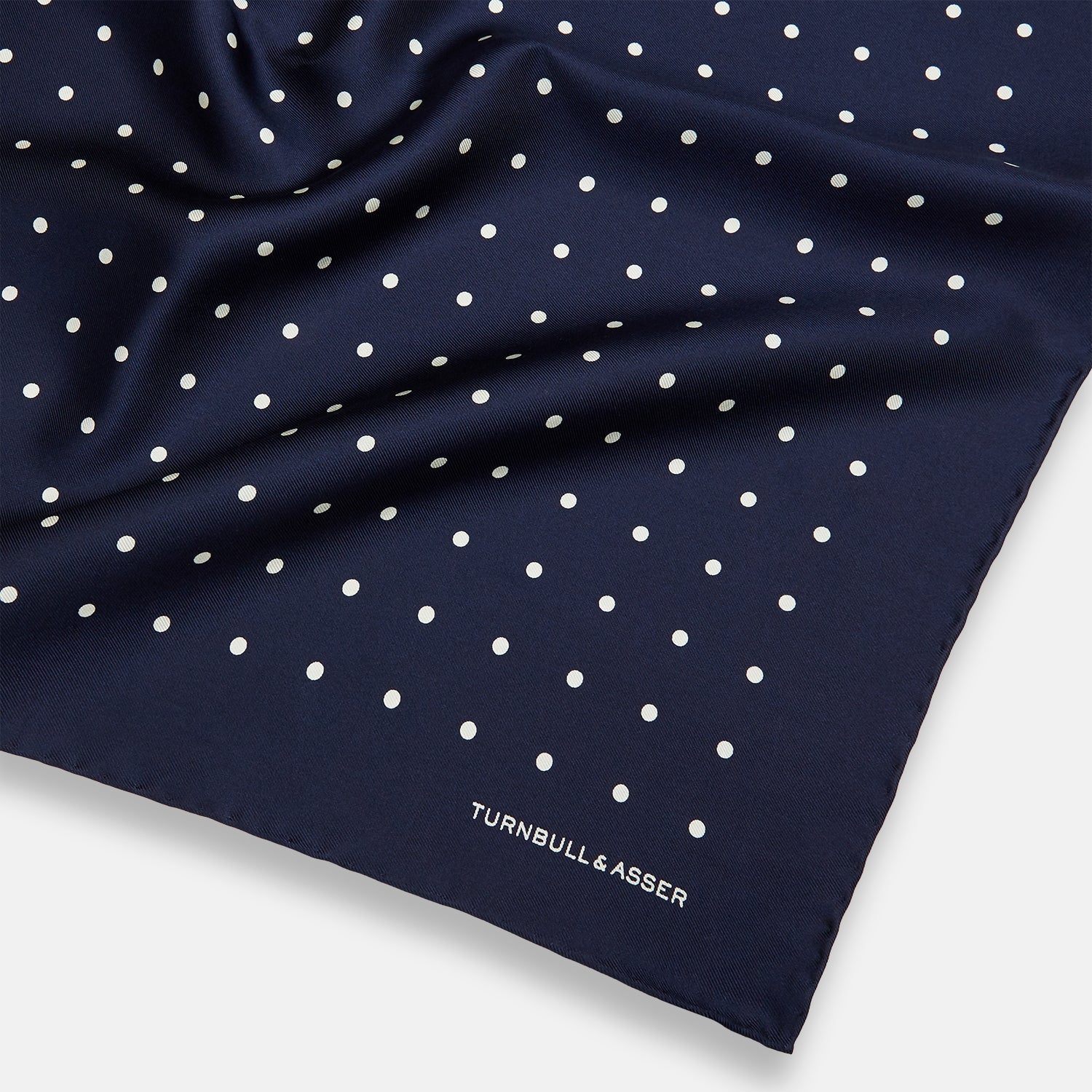 View 2 of product The Turnbull & Asser Navy and White Spot Silk Pocket Square features evenly spaced white polka dots and the brand logo in one corner—a stylish finishing touch for formal looks. One corner is neatly folded over.