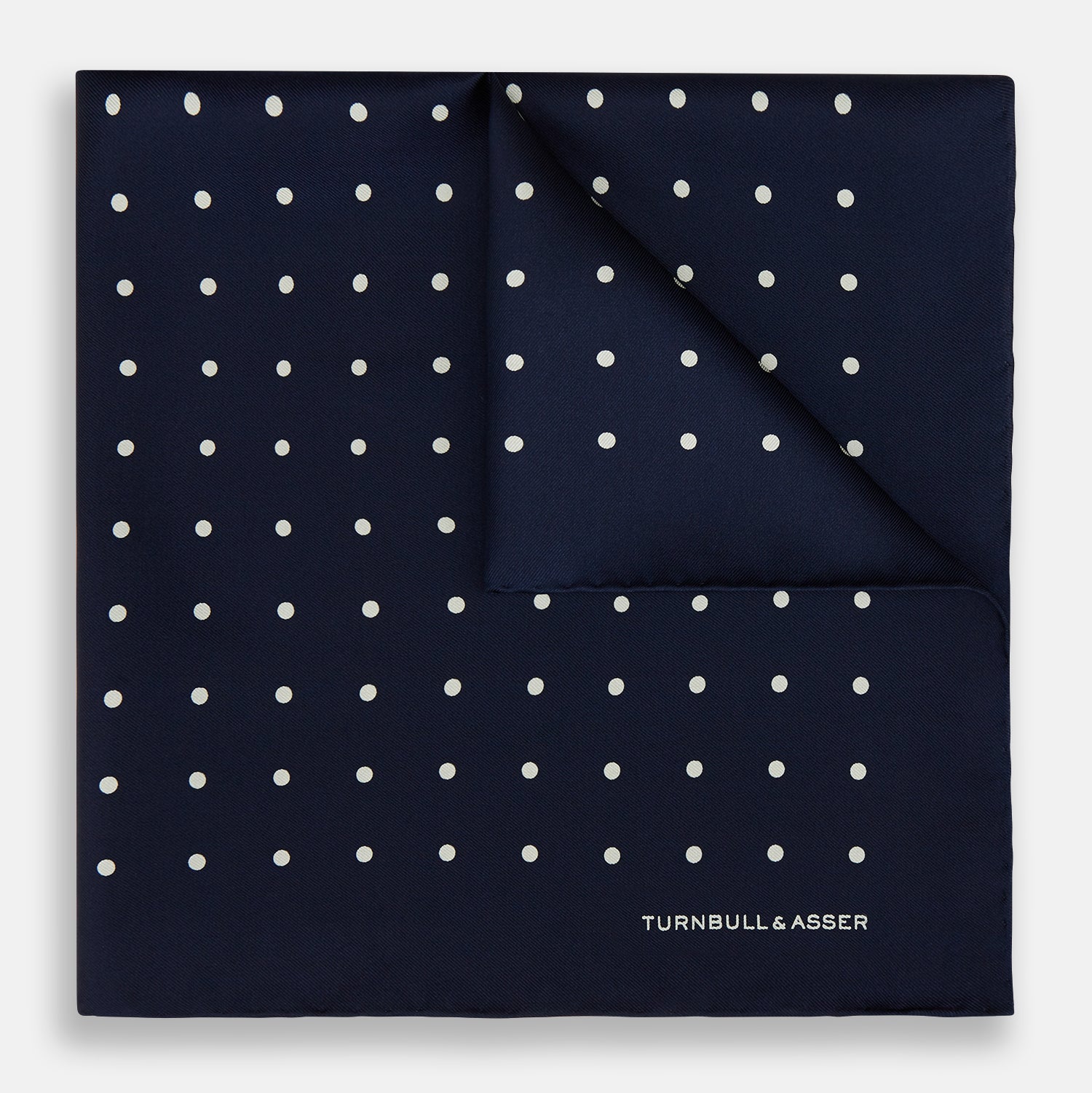 View 1 of product The Turnbull & Asser Navy and White Spot Silk Pocket Square features evenly spaced white polka dots and the brand logo in one corner—a stylish finishing touch for formal looks. One corner is neatly folded over.