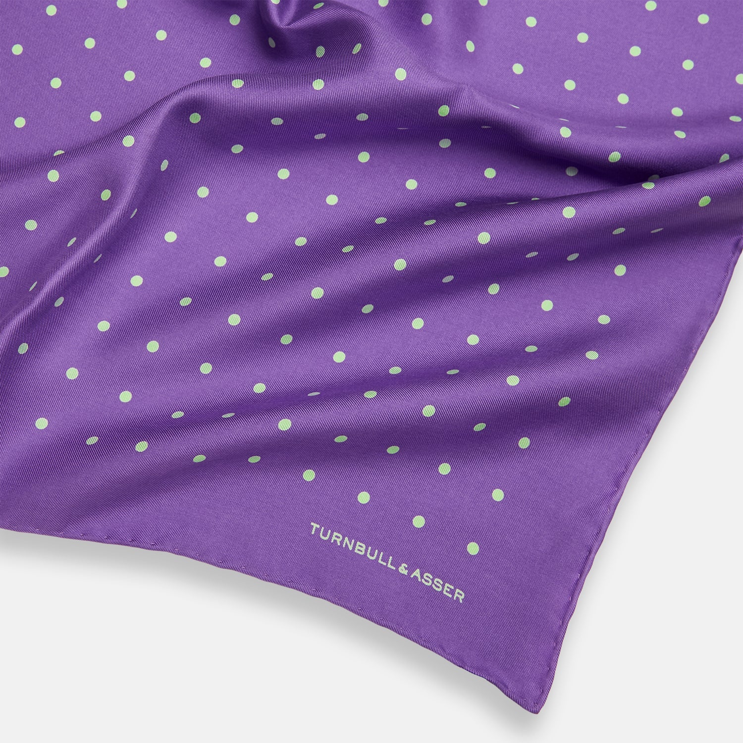View 2 of product The PURPLE AND MINT SPOT SILK POCKET SQUARE by Turnbull & Asser features mint spots on purple silk and the brand's white logo in the lower right corner. One corner is folded over, perfect for elevating any gentleman’s formal look.