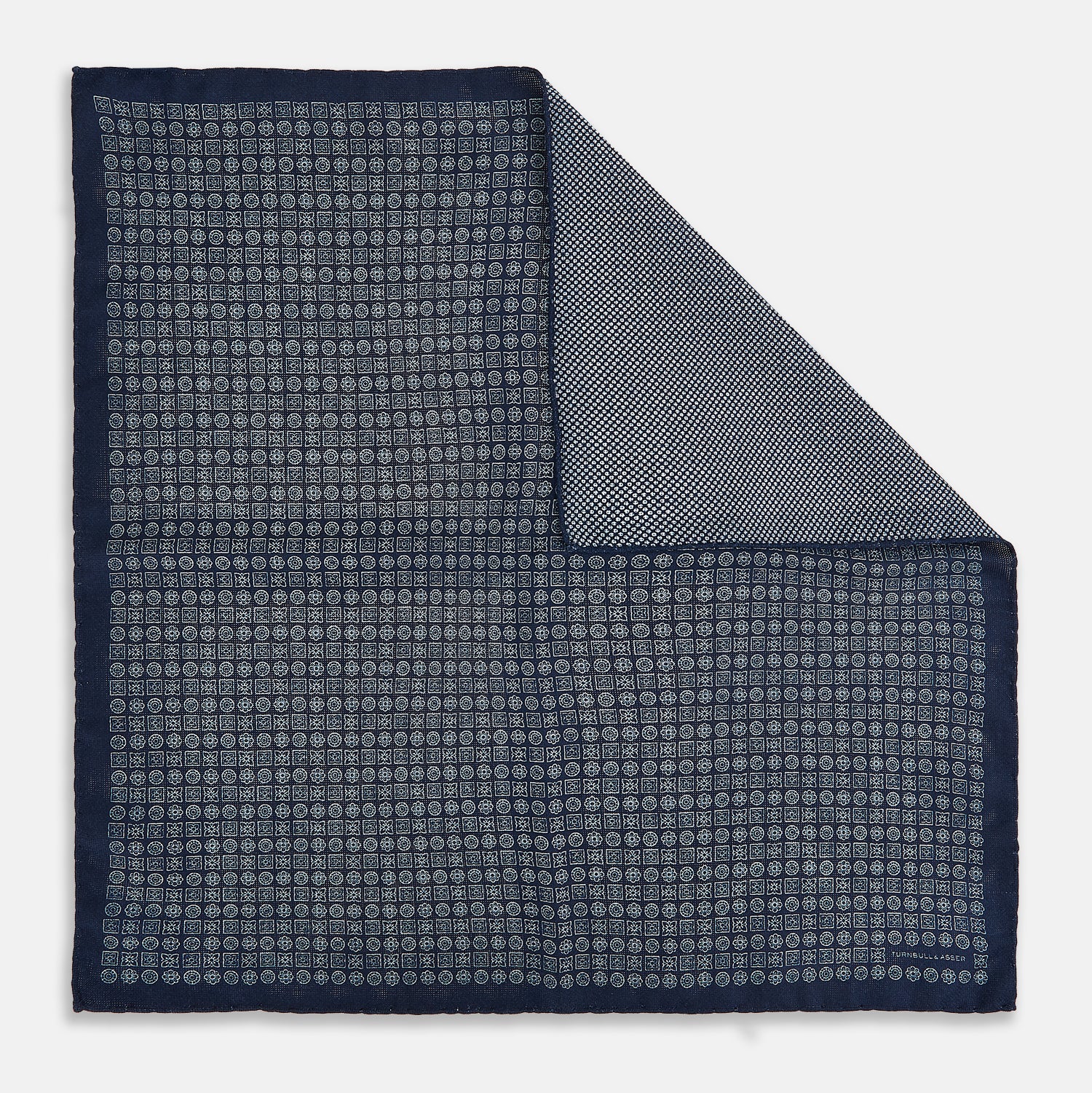 View 1 of product The Rosette Navy Pocket Square in Silk by Turnbull & Asser UK is a square, navy blue accessory featuring a small geometric pattern. Shown laid flat with one corner folded, it reveals a contrasting, finer motif on the reverse side. This elegant double-sided design offers versatile styling options for any occasion.