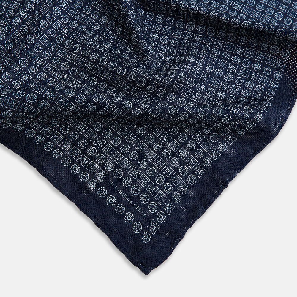 The Rosette Navy Pocket Square in Silk by Turnbull & Asser UK is a square, navy blue accessory featuring a small geometric pattern. Shown laid flat with one corner folded, it reveals a contrasting, finer motif on the reverse side. This elegant double-sided design offers versatile styling options for any occasion.
