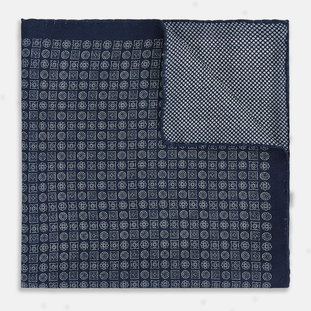 The Rosette Navy Pocket Square in Silk by Turnbull & Asser UK is a square, navy blue accessory featuring a small geometric pattern. Shown laid flat with one corner folded, it reveals a contrasting, finer motif on the reverse side. This elegant double-sided design offers versatile styling options for any occasion.